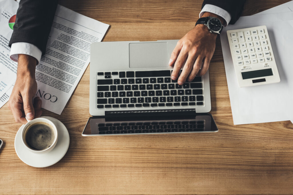 Top view of a person in a suit in front of a desk with a laptop, calculator, documents, and a cup of coffee on it.